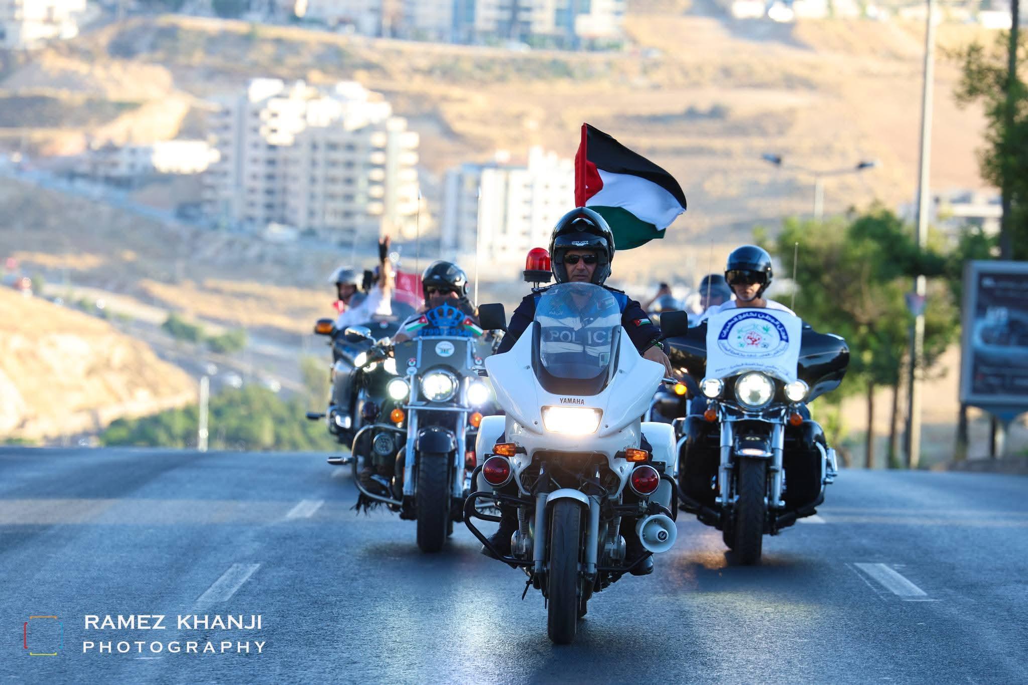 دورية شرطة تتقدّم المسيرة على طريق مرتفع، ودراج خلفها يرفع علمًا أردنيًا كبيرًا فوق أحياء المدينة