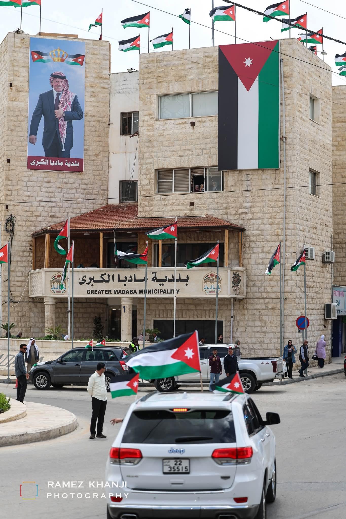 مبنى بلدية مادبا الكبرى تغطّيه الأعلام الأردنية وصورة كبيرة لجلالة الملك، ومركبة من الموكب تمرّ أمامه
