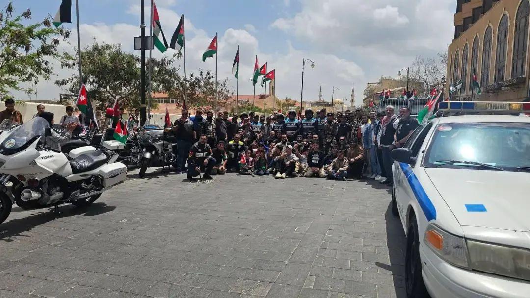 صورة جماعية للدراجين على ساحة مبلّطة بين دراجتي شرطة وسيارة دورية، والأعلام الأردنية ترفرف ومآذن مسجد في الخلفية