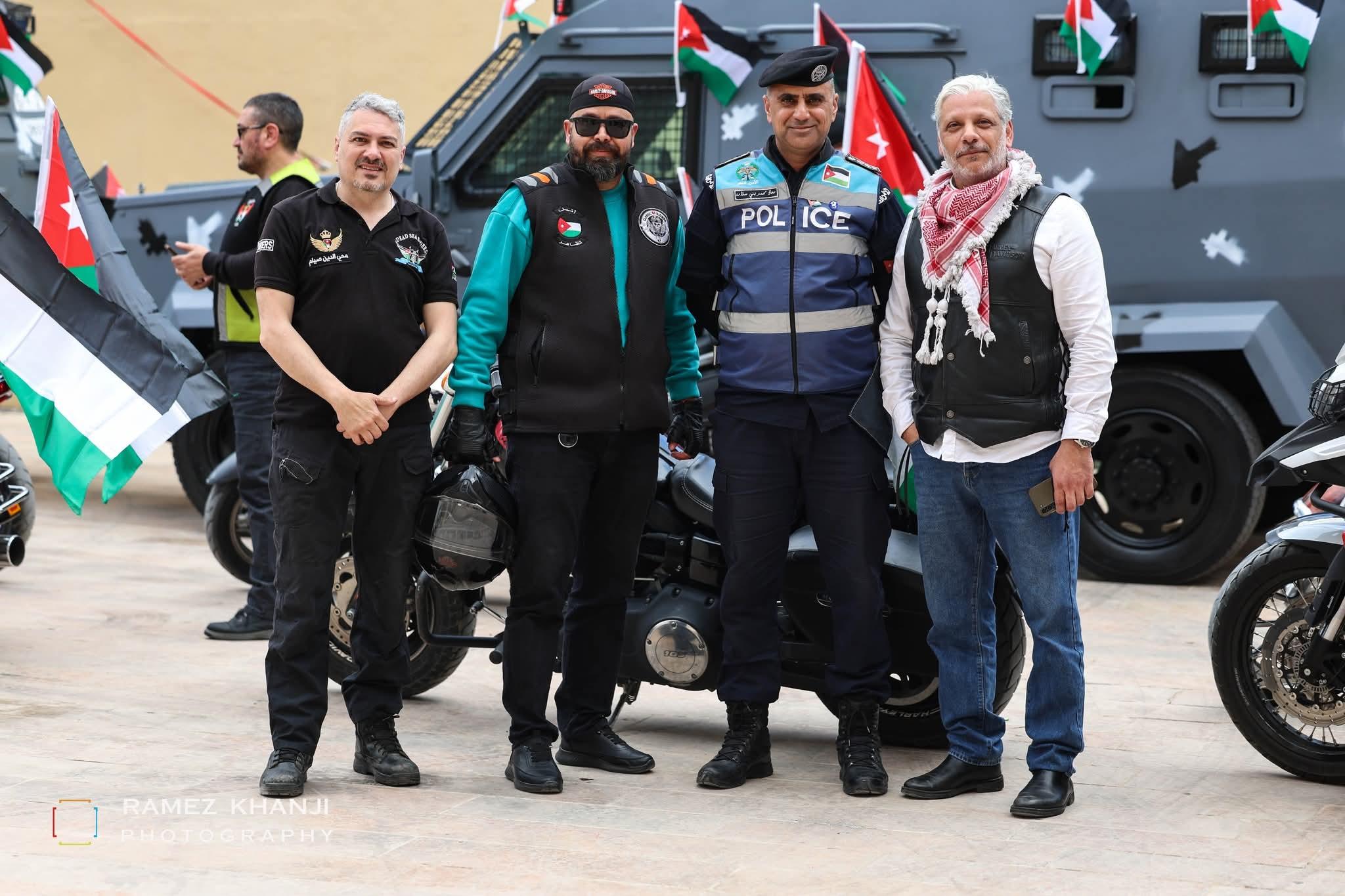 ثلاثة من الدراجين مع ضابط من الأمن العام أمام مركبة مدرّعة تزيّنها الأعلام الأردنية