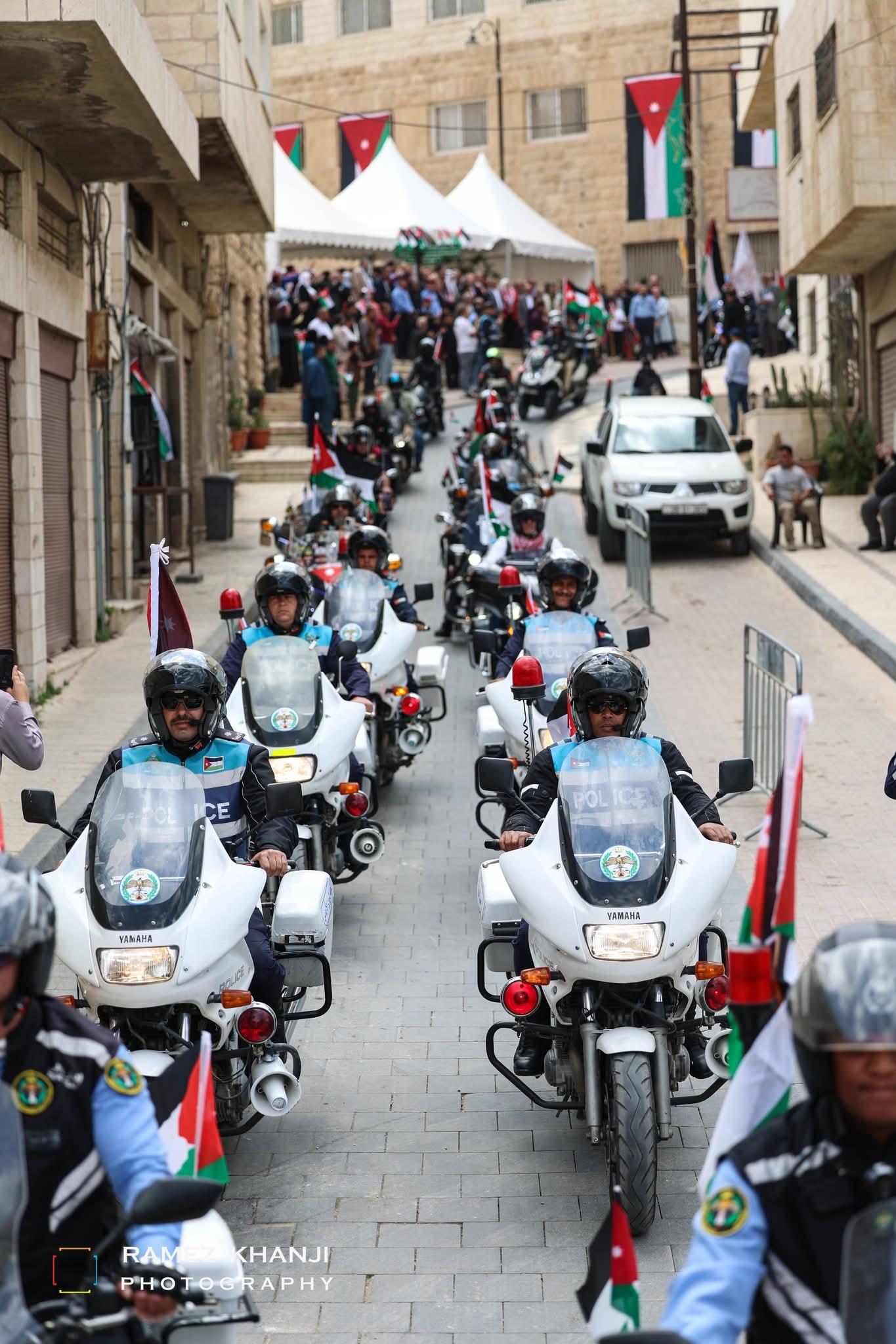 رتلان من دوريات الأمن العام ودراجات القافلة ينزلان شارعًا مبلّطًا بالحجر، والأعلام معلّقة على الشرفات وحشد ينتظر في أعلى الشارع